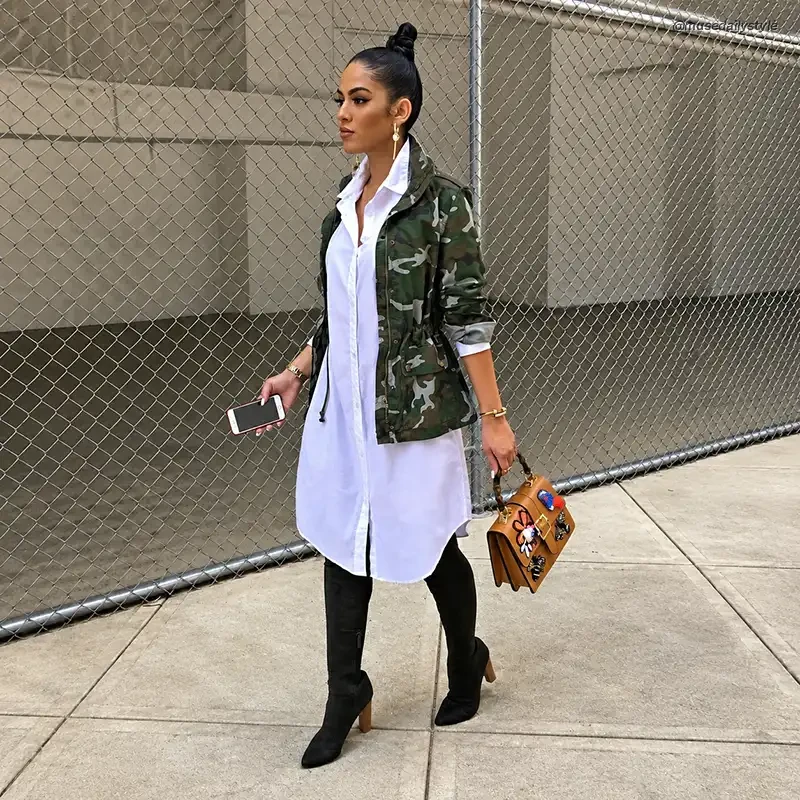 Woman wearing a camo utility jacket over a long white button-down shirt dress with black knee-high boots and a tan top-handle bag