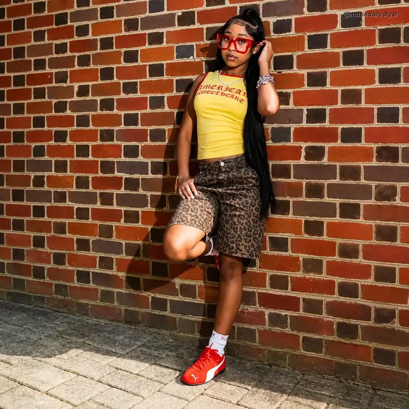 Woman leaning against a brick wall wearing cheetah print shorts, a yellow graphic tank top and red sneakers